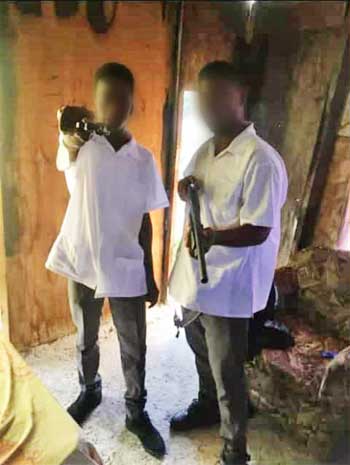 Image: Two boys pictured in full secondary school uniform with what appears to be firearms (a hand gun and shot gun).